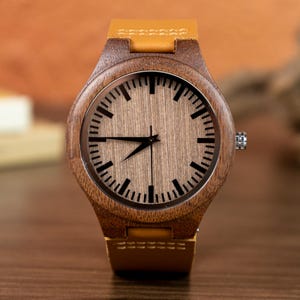 Puede incluir: Un reloj de pulsera de madera con una correa de cuero marr&oacute;n claro. La esfera del reloj es de color marr&oacute;n claro con marcadores de hora y agujas negras. La caja del reloj es de madera marr&oacute;n oscuro.