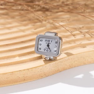 May include: A silver-toned ring watch with a rectangular face. The watch face is white with black numerals and hands, and the brand name "BOZHI" is printed on the face. The ring is set on a light-colored wooden surface.
