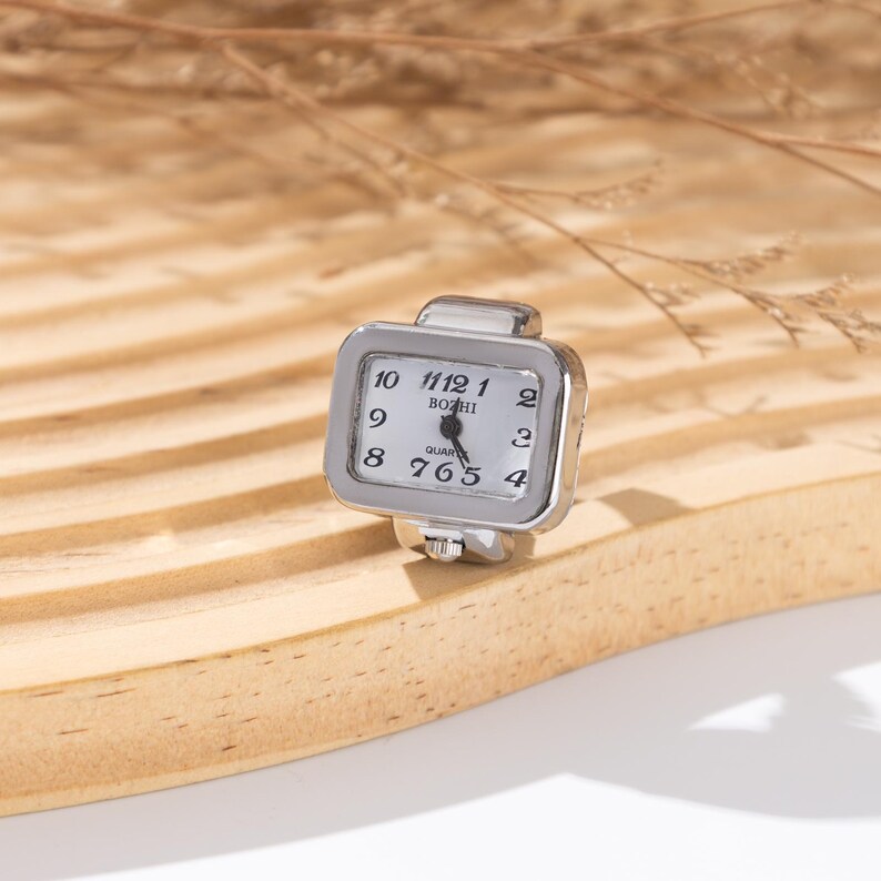 May include: A silver-toned ring watch with a rectangular face. The watch face is white with black numerals and hands, and the brand name "BOZHI" is printed on the face. The ring is set on a light-colored wooden surface.