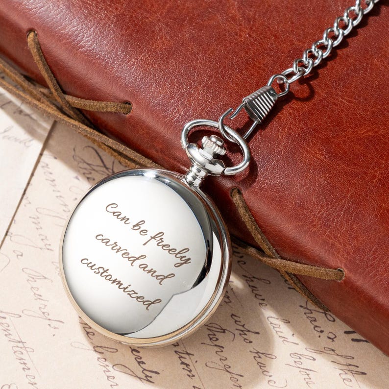 Puede incluir: Un reloj de bolsillo plateado con una cadena, apoyado sobre una superficie de cuero marr&oacute;n. La esfera del reloj tiene el texto "Can be freely carved and customized". El reloj es redondo y tiene una superficie brillante y reflectante.