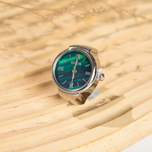 May include: A silver ring watch with a green face and Roman numeral hour markers. The watch face has a marbled appearance. The brand name "BOGU" is printed on the watch face. The watch is set on a light brown wooden surface.