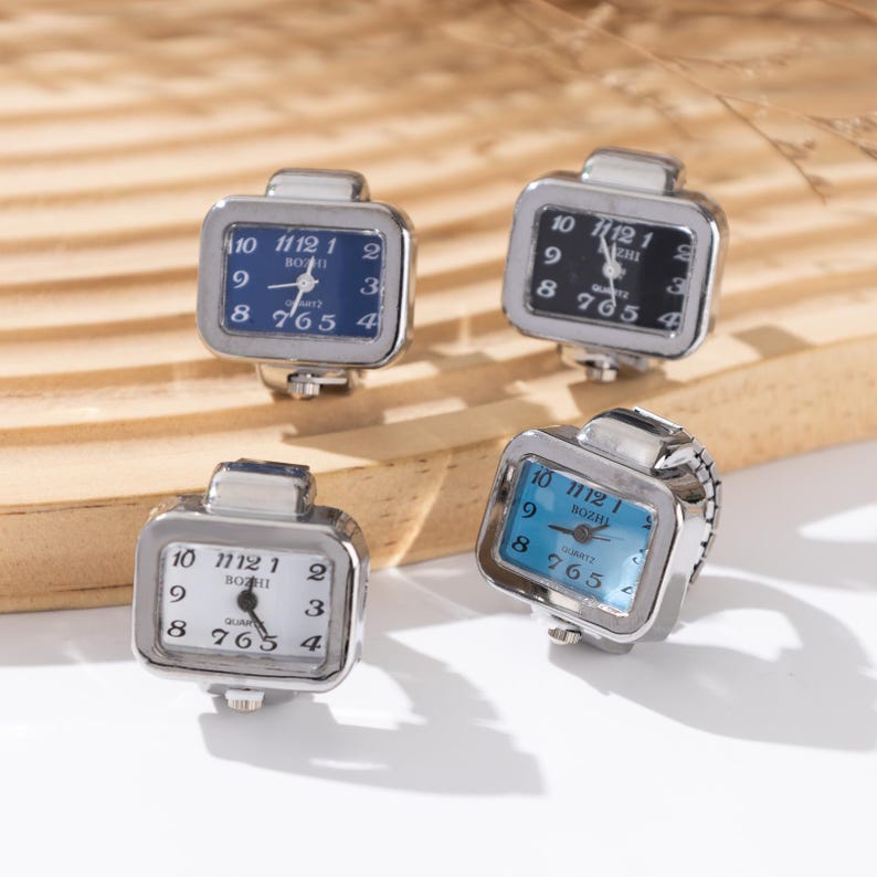 May include: Four square-faced ring watches with silver-toned metal frames. The watch faces have different colored backgrounds: blue, black, white, and light blue. Each watch face displays the time with black numerals and hands.