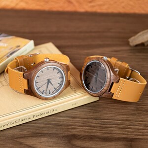Puede incluir: Dos relojes de madera con correas de cuero marr&oacute;n claro. Las esferas de los relojes son de color marr&oacute;n claro con manecillas y marcadores negros. Los relojes est&aacute;n sobre un libro con el texto "Center & Classic Pedestal 34".