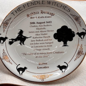 May include: A white decorative plate with a gold rim, featuring black silhouettes of a witch on a broom, black cats, and a tree. Text includes "The Pendle Witches" and historical details. The plate is from the London Heritage Arts & Crafts Centre.