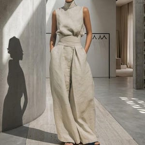 Könnte beinhalten: Beiges Leinen-Outfit, bestehend aus einem ärmellosen Oberteil mit hohem Ausschnitt und einer Hose mit weitem Bein und hohem Bund mit Taschen. Das Outfit wird in einem modernen, minimalistischen Innenraum mit Schatten an der Wand getragen.