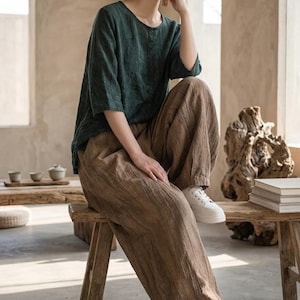 May include: A dark green round-neck top with three-quarter length sleeves paired with loose-fitting brown trousers featuring a textured, crinkled fabric finish and white sneakers. The outfit consists of a simple shirt and wide-leg pants.