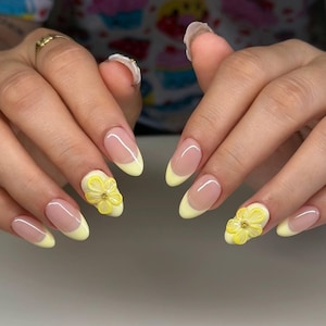 May include: Oval-shaped nails with a French manicure design featuring a pale yellow tip and a clear base. Two nails have 3D yellow flower embellishments. The nails are well-manicured and polished.