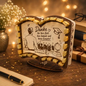 Könnte beinhalten: Ein hölzerner Aufsteller in Buchform mit einer Lasergravur eines Lehrers und Schülern, versehen mit dem Text Danke für Ihre Zeit, Ihre Geduld und Ihren Einsatz! sowie Ihre Abschlussklasse 2026, verziert mit gold verpackten Schokoladenstücken.