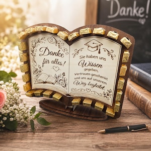 May include: A wooden book-shaped gift with the words "Danke für alles!" and "Sie haben uns Wissen gegeben" engraved on the pages. The edges are lined with gold-wrapped chocolates. A graduation cap is also depicted.