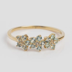 Op de afbeelding: Een goudkleurige ring met drie bloemmotieven. Elk motief bestaat uit kleine, lichtblauwe en lichtroze edelstenen. De ring heeft een dunne, gladde band en de edelstenen zijn dicht op elkaar geplaatst, wat een delicaat en elegant ontwerp creëert.