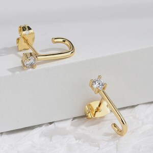 Puede incluir: Un par de pendientes dorados y curvados con una pequeña gema transparente en el extremo de cada barra curva. Los pendientes tienen un cierre de poste y mariposa. Los pendientes se muestran sobre una superficie blanca.