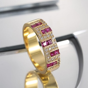 Peut inclure: Une bague en or avec un motif rectangulaire. La bague présente des rangées alternées de pierres roses rectangulaires et de petites pierres claires et rondes. La bague est posée sur une surface réfléchissante.