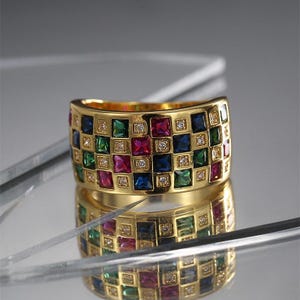 Puede incluir: Un anillo dorado con un diseño de mosaico. El anillo está adornado con piedras cuadradas de colores variados, incluyendo rojo, azul, verde y transparente. Pequeñas piedras transparentes están intercaladas. Se refleja en la superficie.
