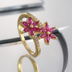 May include: A gold-colored ring featuring two flower designs with vibrant pink petals. The flowers are adorned with small, clear gemstones. The ring's band is also embellished with clear gemstones, creating a sparkling floral jewelry piece.