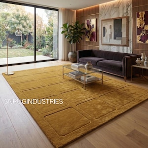 May include: Mustard yellow rectangular area rug with a geometric square pattern. The rug is in a living room setting with a dark brown sofa, glass coffee table, and artwork on the wall. The text "SNRUINDUSTRIES" is visible on the rug.
