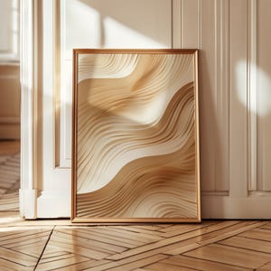 May include: A framed abstract art print featuring a layered design of flowing, wave-like forms in shades of beige and cream. The artwork is encased in a gold-colored frame and leans against a white wall.