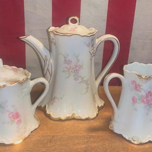 May include: A vintage porcelain tea set featuring a teapot, creamer, and lidded sugar bowl. The white pieces are adorned with pink floral patterns and gold trim. The set is displayed on a wooden surface.
