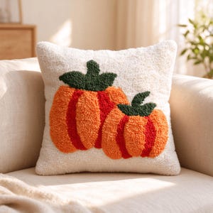 May include: A square throw cushion with a cream-coloured, textured, tufted surface. The front features a design of two pumpkins in vibrant orange and red tones, each topped with dark green stems and leaves, created with a raised, looped yarn texture.