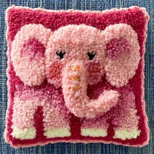May include: A square punch needle pillow featuring a textured pink elephant design. The elephant has large ears, a trunk with orange accents, and closed eyes. The background is a deep magenta, and the elephant's feet are a creamy white color.