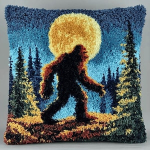 May include: A square throw pillow featuring a textured punch needle design of a dark, hairy creature walking through a forest at night. A large, bright yellow full moon illuminates the scene, with deep blue sky and green pine trees in the background.