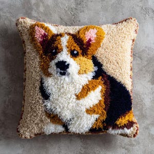 Puede incluir: Cojín decorativo cuadrado con un diseño texturizado de un perro Corgi. La cara del perro está representada en blanco, marrón, negro y amarillo sobre un fondo beige. Las orejas del perro son rosas. Un elemento decorativo para el hogar.