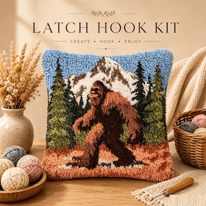 Könnte beinhalten: Ein quadratisches Knüpfkissen-Set mit einer texturierten Bigfoot-Figur, die durch einen Wald vor schneebedeckten Bergen läuft. Der Text LATCH HOOK KIT CREATE HOOK ENJOY ist in einer dunkelbraunen Schrift über dem Kissen abgedruckt.