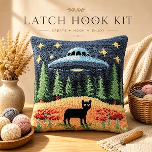 May include: A square latch hook kit cushion featuring a dark blue night sky with yellow stars and a blue UFO hovering over a forest of green pine trees. A black cat stands in a tan field with red bushes. Text reads LATCH HOOK KIT CREATE HOOK ENJOY.
