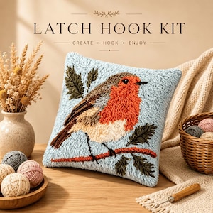 Op de afbeelding: Een vierkant knooppakket kussen met een getextureerd roodborstje op een lichtblauwe achtergrond, zittend op een tak met groene bladeren. De tekst LATCH HOOK KIT CREATE HOOK ENJOY staat boven het kussen, vergezeld door bollen wol.