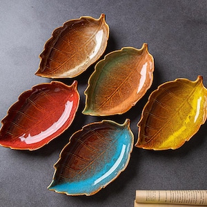 Puede incluir: Seis platos de cerámica con forma de hoja en tonos rojos, marrones, verdes y amarillos. Cada plato tiene un acabado brillante y patrones de vetas detallados. Los platos están dispuestos sobre una superficie gris oscuro, mostrando sus colores y formas únicos.