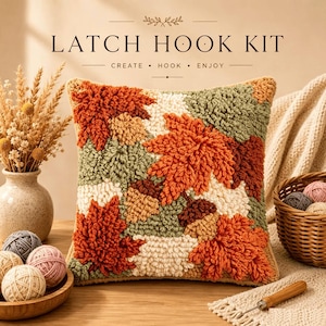 Peut inclure: Un coussin de kit de crochet à loquet carré avec un motif texturé de feuilles d'automne et de glands dans des tons orange, crème et vert sauge. Le texte LATCH HOOK KIT CREATE HOOK ENJOY est imprimé au-dessus du coussin entouré de pelotes.