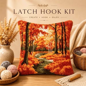 May include: A square latch hook kit pillow featuring a vibrant autumn forest scene with orange and red foliage, dark tree trunks, and a winding path. The text LATCH HOOK KIT CREATE HOOK ENJOY is printed above the pillow in a dark brown font.