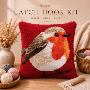 Peut inclure: Un coussin de kit de crochet à loquet carré avec un fond rouge et un motif central de rouge-gorge dans des tons orange, blanc et marron. Le texte LATCH HOOK KIT CREATE HOOK ENJOY est imprimé au-dessus, avec des pelotes de laine.