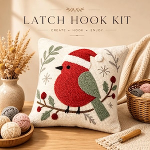 Peut inclure: Un kit de crochet à loquet comprenant un coussin carré avec un oiseau rouge portant un bonnet de Père Noël, perché sur une branche avec des baies rouges et des feuilles vertes. Le texte LATCH HOOK KIT CREATE HOOK ENJOY est visible au-dessus.
