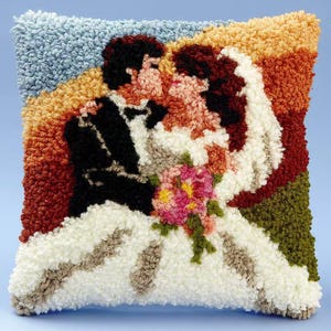 May include: A square punch needle throw pillow featuring a textured, tufted design of a couple in wedding attire. The figures are depicted in black and white against a background of blue, orange, and green, with a pink floral bouquet in the center.