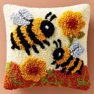 May include: A square throw pillow featuring a textured punch needle design on a cream background. The design displays two bees with black and yellow striped bodies and light gray wings, surrounded by orange and yellow flowers with green stems.