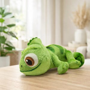 Peut inclure: Une peluche verte en forme de caméléon avec une expression amicale. Le jouet a un corps vert clair avec des rayures vertes plus foncées et une queue enroulée. Il présente un œil marron et est posé sur une surface en bois.