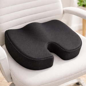 May include: A black ergonomic seat cushion with a contoured design, placed on a white office chair. The cushion has a U-shaped cutout in the front and a textured surface, designed for comfort and support.