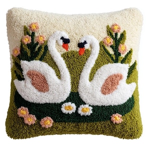 May include: A square throw cushion featuring a textured punch needle design of two white swans facing each other. The swans are set against a cream background with green foliage, pink flowers, and two white daisies at the bottom of the scene.