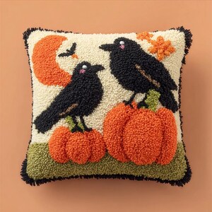 Op de afbeelding: Een vierkant punch needle sierkussen met een crèmekleurige achtergrond, twee zwarte kraaien op oranje pompoenen. Een oranje maan en kleine oranje blaadjes zijn bovenaan zichtbaar, met een groene basis en zwarte franje aan de rand.