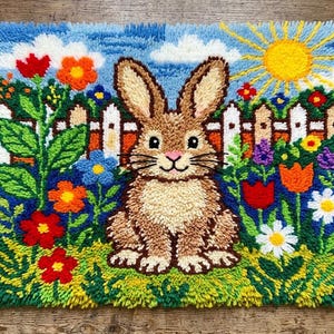 Op de afbeelding: Een rechthoekig knoopkleed met een bruin konijn dat in een tuin zit. De scène bevat een wit tuinhek, een heldergele zon in een blauwe lucht met witte wolken en diverse kleurrijke bloemen in rood, oranje, blauw en wit.