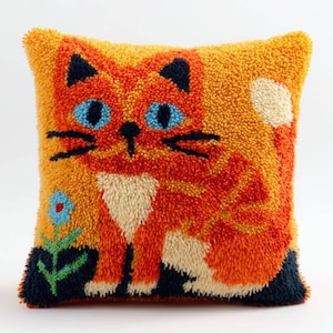 Op de afbeelding: Vierkantig kussen in oranje en geel met een kattenontwerp. De kat heeft blauwe ogen, zwarte oren en snorharen en een witte borst. Een blauwe bloem staat linksonder. Het kussen is gemaakt van een textuurrijk materiaal.