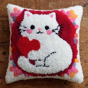 Peut inclure: Un coussin carré représentant un chat blanc aux joues roses tenant un cœur rouge. Le chat est centré sur un fond rouge profond, entouré d'une bordure de petites formes florales multicolores dans des tons de rose, d'orange et de violet.