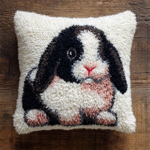 Peut inclure: Un coussin carré blanc cassé avec une texture touffue. Le coussin présente une image détaillée d'un lapin avec des oreilles noires et marron, un œil noir et un nez rose. Le corps du lapin est un mélange de blanc, de rose et de marron.