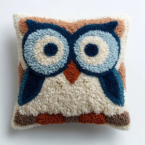 May include: Square cushion with a detailed owl design. The owl has large blue eyes, a rust-coloured beak, and a cream-coloured body. The cushion's border is brown and tan, creating a textured, plush appearance.