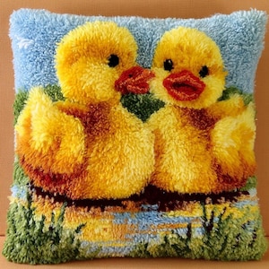 May include: A square latch hook pillow featuring two fluffy yellow ducklings with orange beaks. The ducks are positioned against a light blue sky background with green grass and a small pond at the bottom, rendered in a textured, looped yarn pile.