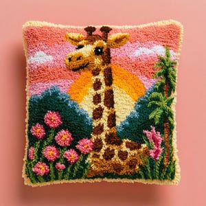 Peut inclure: Un coussin carré avec un motif en punch needle texturé représentant une girafe sur un fond de coucher de soleil. La scène comprend un feuillage vert foncé, des fleurs rose vif et un palmier, le tout réalisé en boucles de laine.