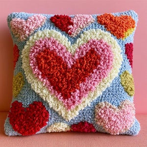 Peut inclure: Un coussin carré à fond bleu clair orné d'un grand cœur central composé de couches concentriques rouges, roses et crème. La surface est recouverte de fils bouclés texturés, avec d'autres cœurs plus petits rouges, roses, orange et jaunes dispersés sur le coussin.