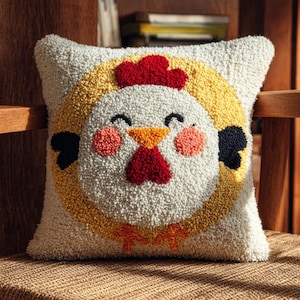 May include: A square throw cushion featuring a textured punch needle design of a chicken face. The chicken has a white face, a yellow circular border, a red comb and wattle, an orange beak, pink cheeks, and black eyes and wing details.