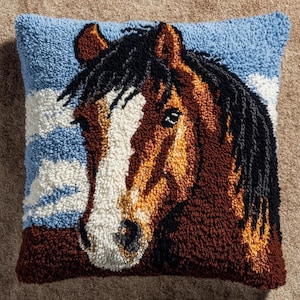 Peut inclure: Un coussin carré avec un motif en punch needle texturé représentant une tête de cheval. Le cheval a un pelage marron, une liste blanche sur le visage et une crinière noire. Le fond montre un ciel bleu clair avec des nuages blancs et un sol marron.