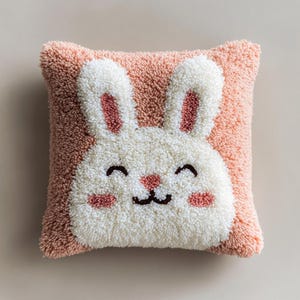 Può includere: Un cuscino quadrato e strutturato con la faccia di un coniglietto sorridente. Il coniglietto ha pelliccia bianca, orecchie e guance rosa e bocca e occhi neri. Lo sfondo del cuscino è rosa chiaro.